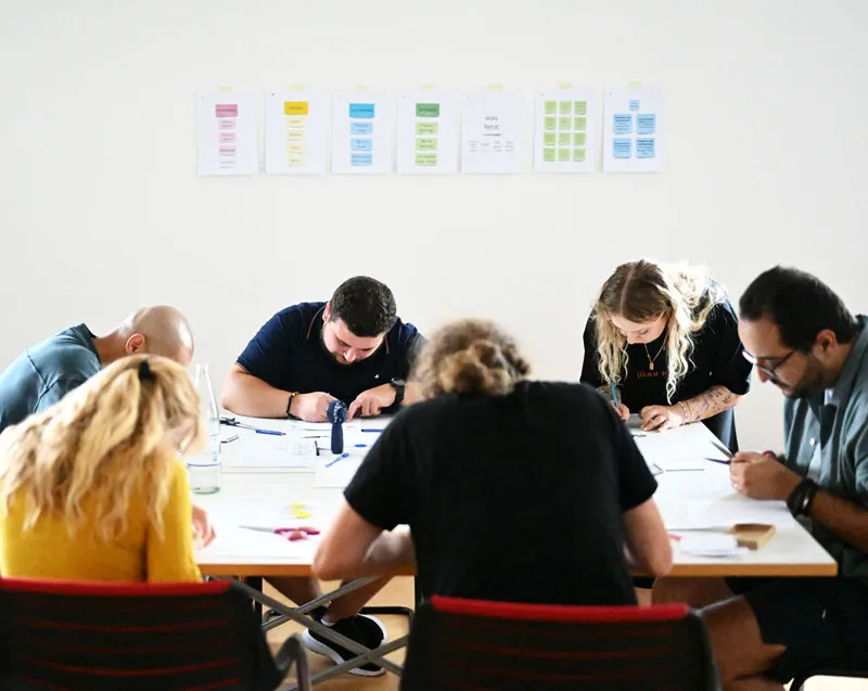 Workshop Impression: Die ganze Gruppe beugt sich an einem Tisch sitzend über die gemeinsame Arbeit.